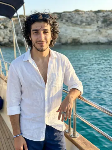 Andreas, AI character: the stern deck of a white charter yacht anchored at the Blue Lagoon on the Akamas coast, turquoise water…