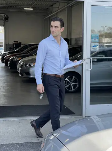 Domingo, AI character: the dealership service entrance in mid-morning, between the glass office door and a line of cars waiting…