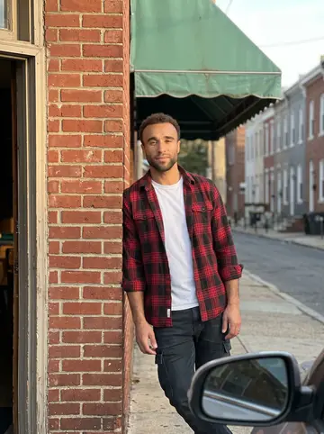 Asher, AI character: on the sidewalk of East Passyunk Avenue in South Philadelphia, leaning on the brick wall beside the front…