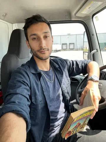 Joel, AI character: the cab of a parked refrigerated truck during a dead hour behind the warehouse, old paperback western and…