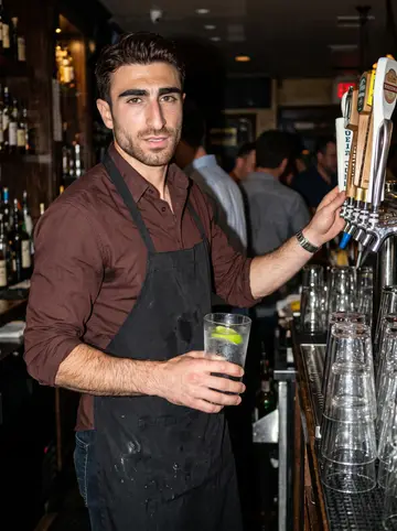 Conor, AI character: inside the pub on a crowded summer night, behind the bar with taps, stacked pint glasses, and regulars…