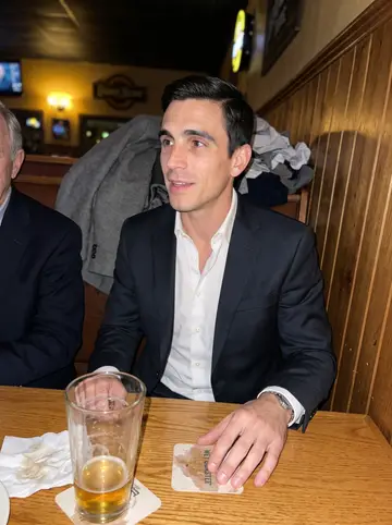 Adam, AI character: a low-key Cleveland neighborhood bar after a drivers' meeting, wood panel wall and half-empty pint glasses…
