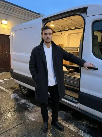Orion, AI character: beside the catering company's white delivery van outside a funeral hall in South Boston, pavement wet from…