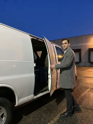 Zackary, AI character: the wet parking lot behind the linen plant before dawn, beside a plain white service van with loading…