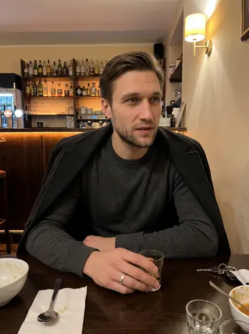 Yevhen, AI character: a narrow Ukrainian bar in Vienna after a drivers' meeting, dark wood table, soup bowls and small glasses…