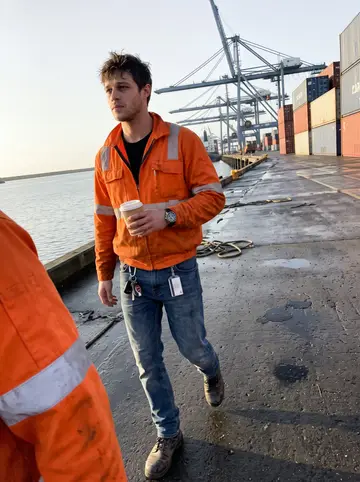 Olivier, AI character: the quay at the Grand Port Maritime de Marseille at the end of his night shift, stacked containers and a…