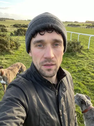 Ronan, AI character: open grass of the Curragh plains at dawn, gorse bushes and a white running rail fading into fog
