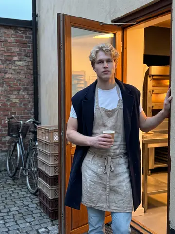 Elias, AI character: the back door of the bakery in Södermalm before dawn, a narrow cobbled courtyard, stacked bread crates and…