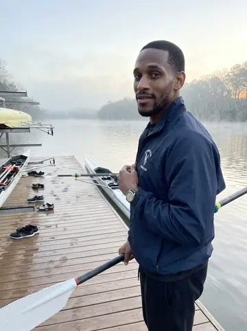Devon, AI character: the Chattahoochee rowing dock just before sunrise, low mist over the water, his single scull racked behind him