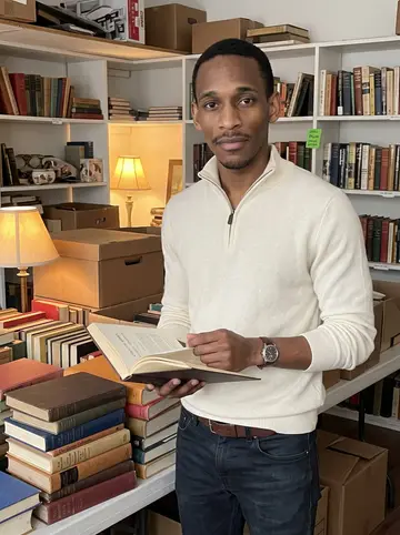 Devon, AI character: a crowded estate sale room in southwest Atlanta, folding tables stacked with old hardcovers and cardboard…