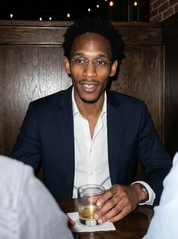 Devon, AI character: a low-lit Edgewood bar after work, two friends cropped at the edges of the booth, dark wood wall behind him