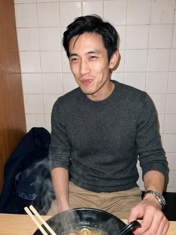 Kenta, AI character: a small Queens ramen booth with a black ramen bowl steaming on the table and a plain tiled wall behind