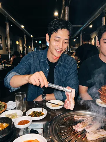Aiden, AI character: a crowded late-night Korean barbecue place in Koreatown with his younger brother, grill smoke and side…