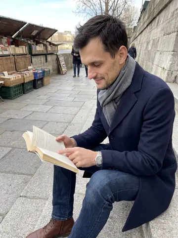 Sébastien, AI character: stone steps near the bouquinistes by the river, a used paperback open in his hands