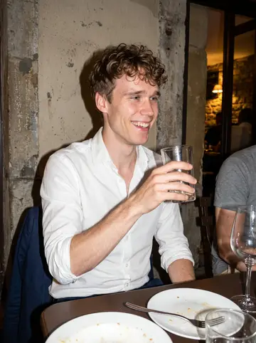 Jean-Baptiste, AI character: a small crowded bar terrace in Lyon at night, friends cropped around the table, empty plates and glasses…
