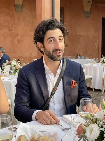 Pietro, AI character: a summer wedding courtyard outside Bologna, seated at table 14 with white tablecloths, flowers, and guests…