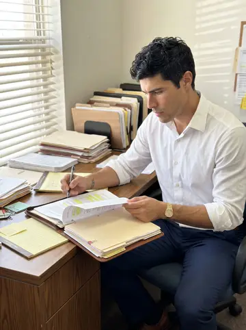 Darío, AI character: his small immigration law office near downtown El Paso, case files stacked on a wood desk, city light…