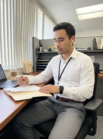 Ricardo, AI character: a small property insurance field office in San Antonio, at a desk with claim folders, a laptop, and a…