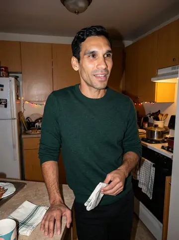 Yunior, AI character: his grandmother's apartment kitchen after Sunday dinner, warm cabinets and a pot on the stove behind him