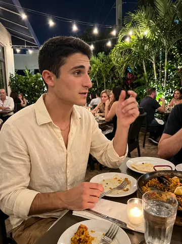 Néstor, AI character: a Spanish restaurant patio in Coral Gables on Sunday night, a paella pan and water glasses on the table