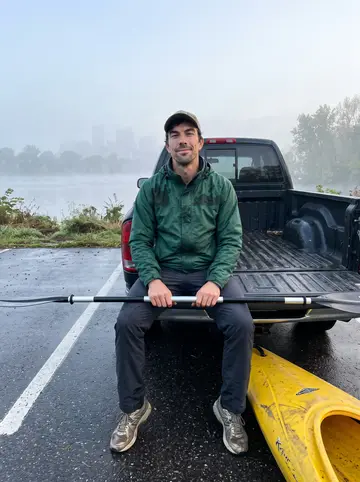 Justin, AI character: Mississippi River launch at dawn beside his battered yellow kayak and a wet parking lot, Minneapolis…