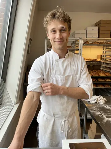 Jacob, AI character: the front window of his Baltimore bakery just after sunrise, trays of croissants cooling behind him
