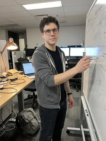Zane, AI character: his logistics startup office in Seattle, standing beside a whiteboard and a messy standing desk after most…