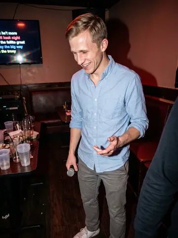 Larry, AI character: a basement karaoke bar in Allston on a humid summer night, crowded table behind him