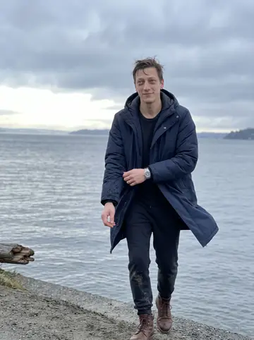 Jeremiah, AI character: Alki Beach just after dawn, grey Puget Sound behind him and low clouds over the water