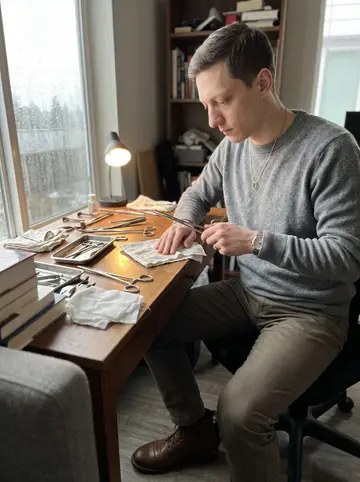 Jeremiah, AI character: his Seattle apartment desk by a rainy window, antique surgical instruments laid out on a cotton cloth…