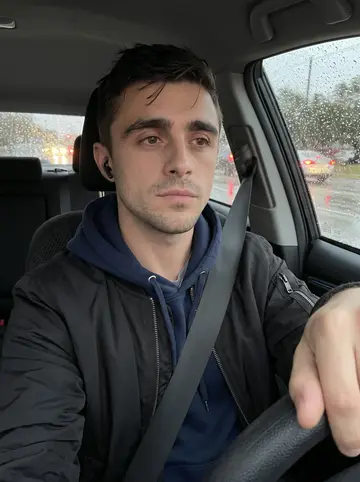 Kobe, AI character: parked in his car after work in Austin drizzle, windshield dotted with rain, dashboard maps glowing faintly