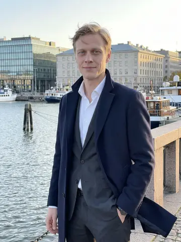 Niko, AI character: Helsinki waterfront near a glass office building at low autumn sun, with the harbor and pale stone…