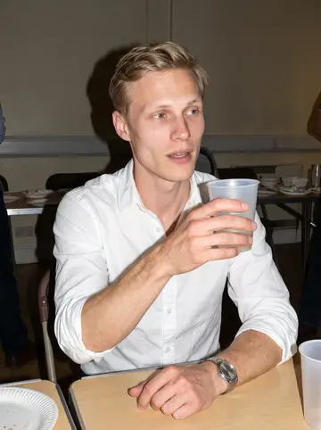 Mykola, AI character: small Ukrainian community fundraiser in a church hall near Cambridge, folding tables and paper plates…