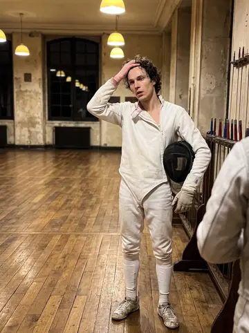 Lucien, AI character: an old salle d'armes in the Marais after midnight, worn wooden floor, a rack of épées, tall dark windows
