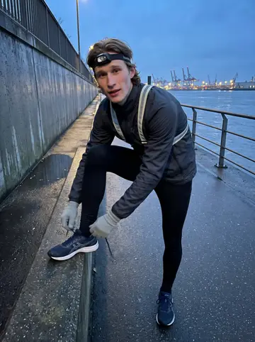 Linus, AI character: on the Elbe river path in Hamburg just after dusk, damp pavement and harbor lights in the distance, his…
