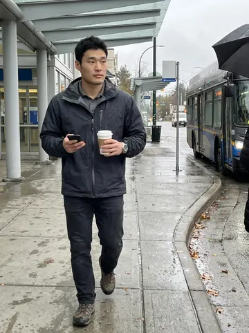 Gordon, AI character: covered sidewalk near a Vancouver SkyTrain station after an early pharmacy shift, wet pavement and buses…