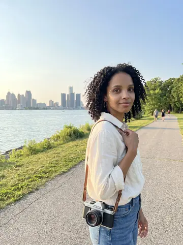 Brianna, AI character: Belle Isle near the Detroit River at golden hour, city skyline soft behind her, her vintage Pentax hanging…
