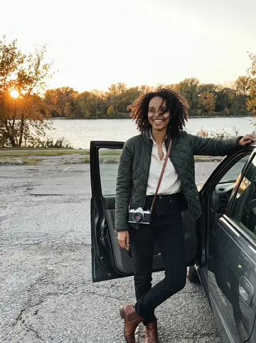 Brianna, AI character: a parking pull-off near Belle Isle at sunset, older dark sedan door open, Detroit River trees turning gold…