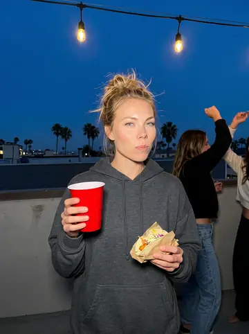 Lena, AI character: a fish taco shop's outdoor patio in Ocean Beach at dusk, paper-lined baskets on a picnic table, strings of…