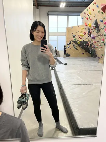 Lin, AI character: a bouldering gym in Dogpatch at 6:30 in the morning, padded floor, colorful holds on the wall behind her,…