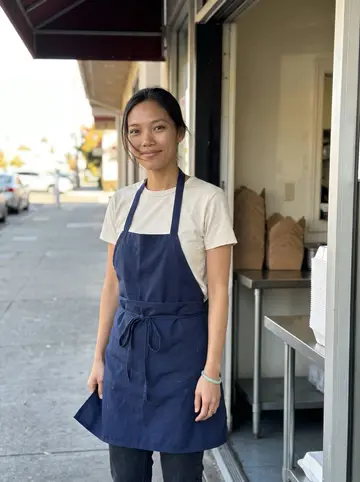 Uyen, AI character: outside her mother's small pho shop in San Jose after the lunch rush, standing in the open side doorway…