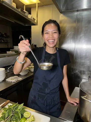 Uyen, AI character: inside the pho shop kitchen late after closing, stainless counter crowded with herbs, limes and a steaming…