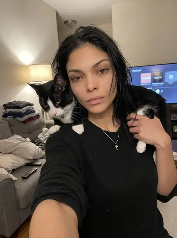 Keila, AI character: her Bronx apartment couch at night, folded laundry and a true-crime podcast paused on the TV behind her