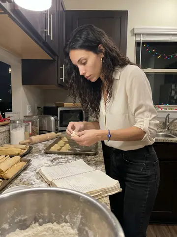 Nadia, AI character: her condo kitchen in Houston, the battered family recipe notebook open beside trays of ma'amoul and tamales