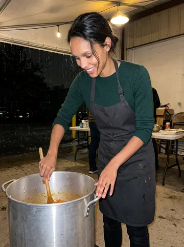 Kemi, AI character: covered warehouse lot during her first-Friday jollof rice cookout for the crew, drizzle outside the awning