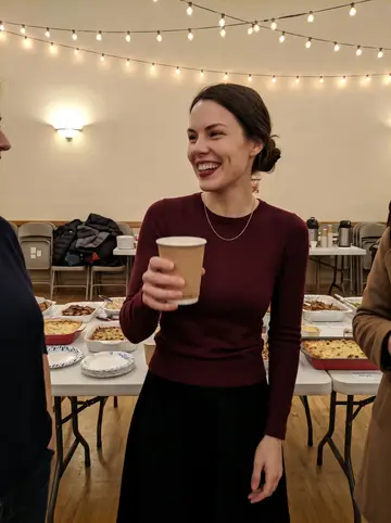 Lyudmila, AI character: community hall holiday potluck in Parma, folding tables with home-cooked dishes and string lights along…