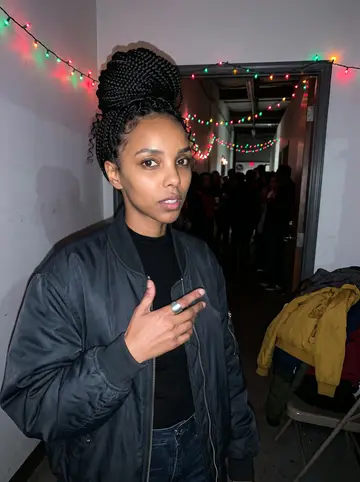 Gifty, AI character: crowded warehouse hallway outside a small Atlanta dance party in December, colored string lights and coats…