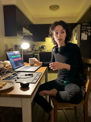 Romy, AI character: her kitchen table late at night, laptop open to a podcast edit and a cheap desk lamp beside a soldering mat