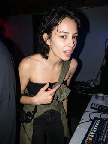 Romy, AI character: a small Pittsburgh basement electronic show after midnight, black walls, people cropped at the edges, a…