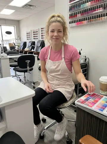 Sandra, AI character: the break corner of a small Milwaukee nail salon, polish racks and a white manicure table behind her